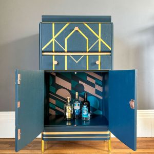 Art Deco cocktail cabinet with petrol blue finish, gold geometric design and patterned interior wallpaper.