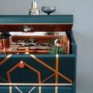 Art Deco cocktail cabinet slightly open to reveal a copper interior with mirrored shelf and bar accessories.